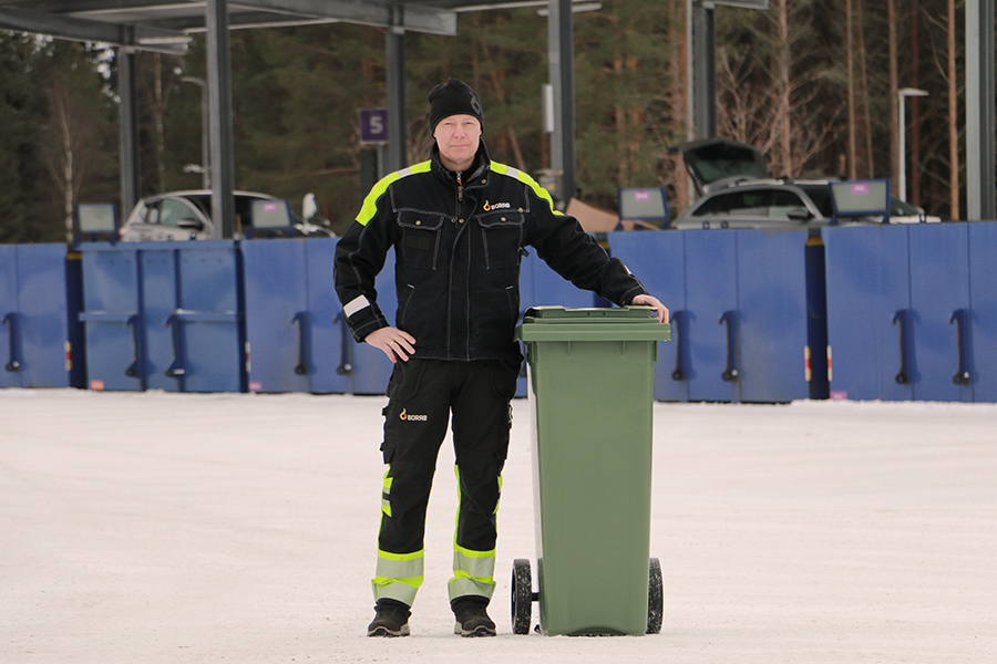 Vår arbetsledare Jörgen står vid ett grönt kärl, i bakgrunden syns återvinningscentralen.